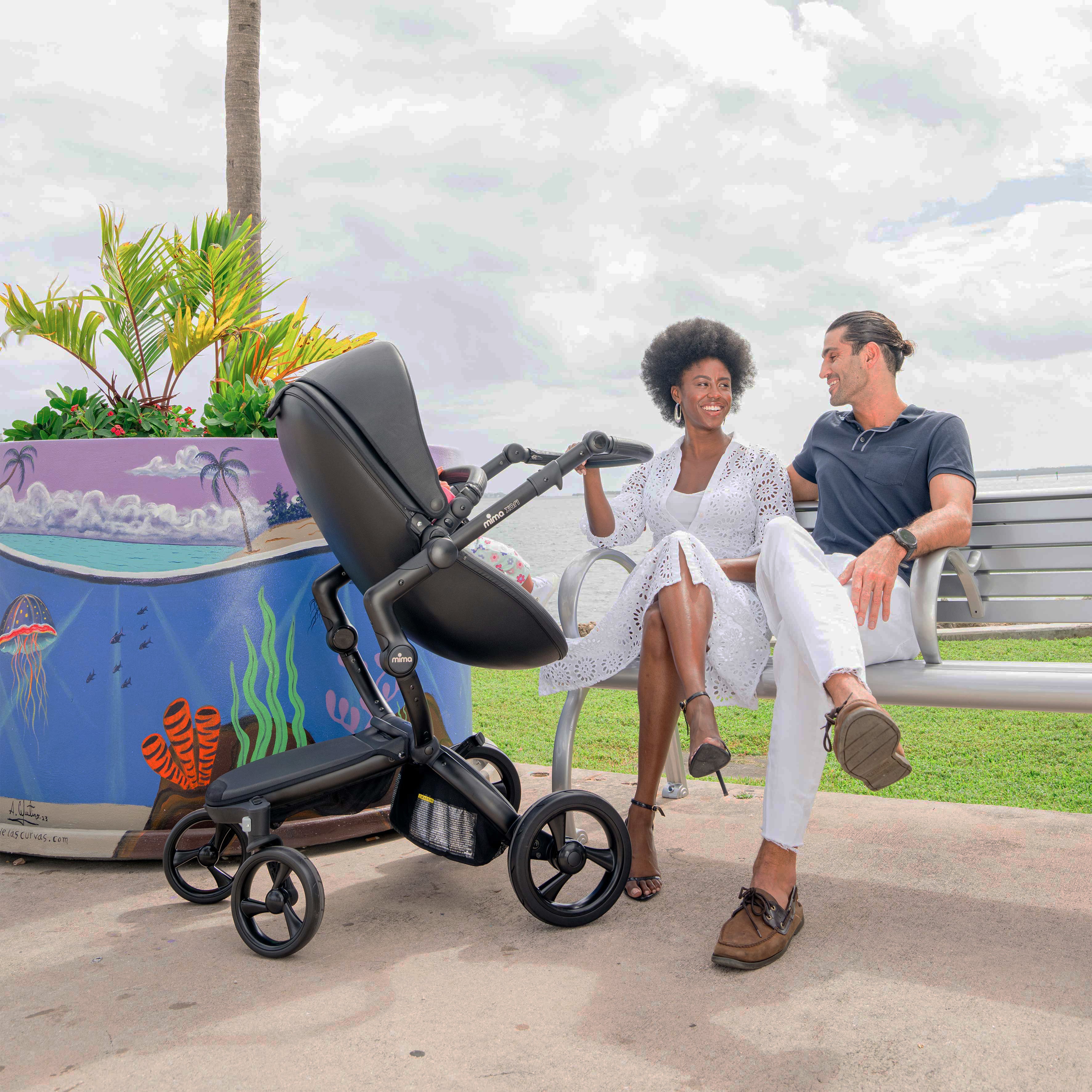 Mima Xari Black stroller on a scenic dock with a couple relaxing on a bench, featuring lush greenery and a colorful ocean-themed planter in the background.