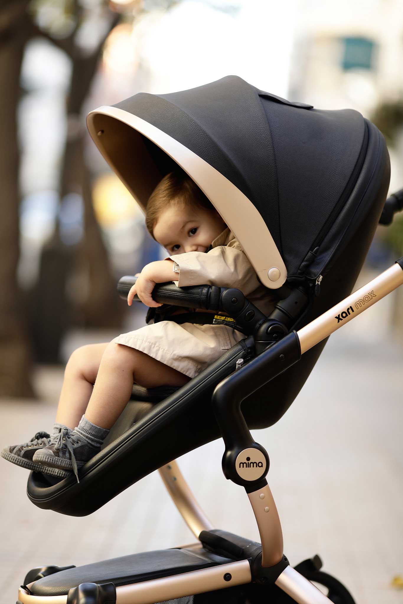 Mima Xari Max Black and Gold Edition stroller on a city street, featuring a child seated in the stroller with a blurred urban background of trees and buildings.