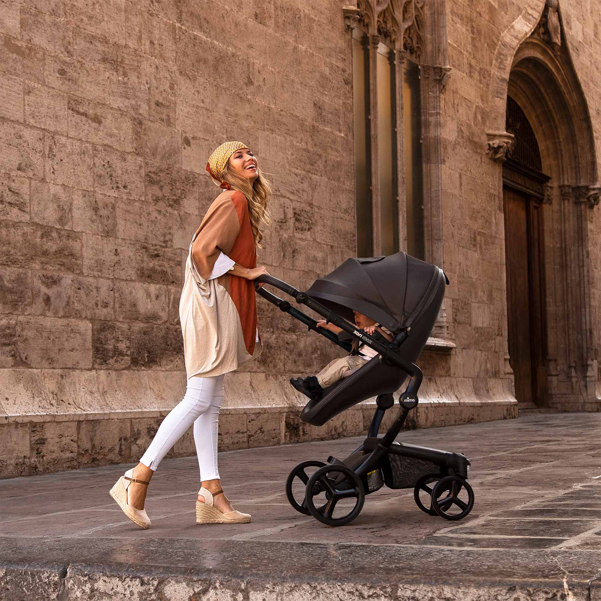 Black mima Xari Max stroller with baby, mother in beige hat pushing, side view on street
