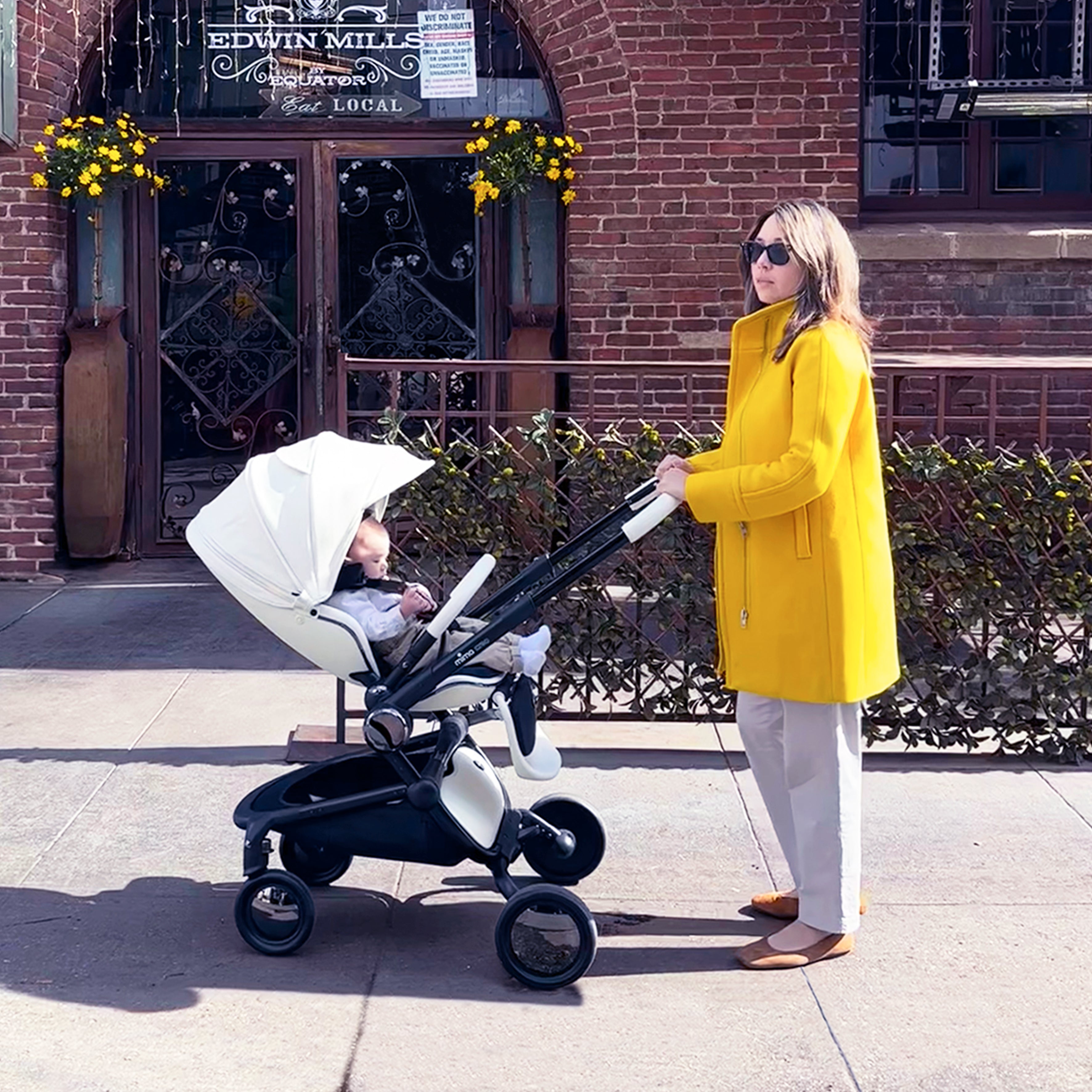 White mima creo stroller with baby, mother in yellow coat pushing, side view on street