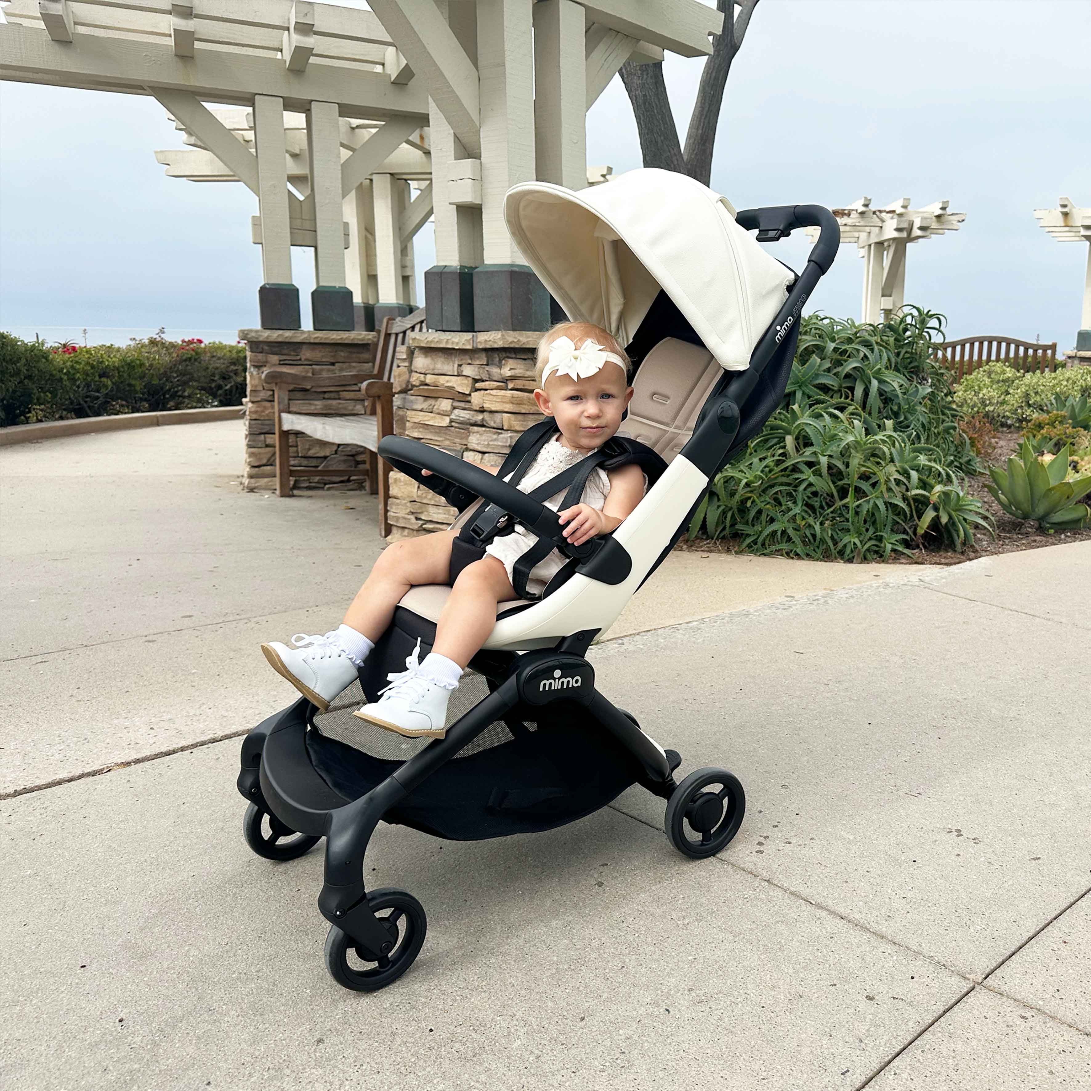 toddler girl sitting in mima miro compact travel stroller in white on a paved coastal path