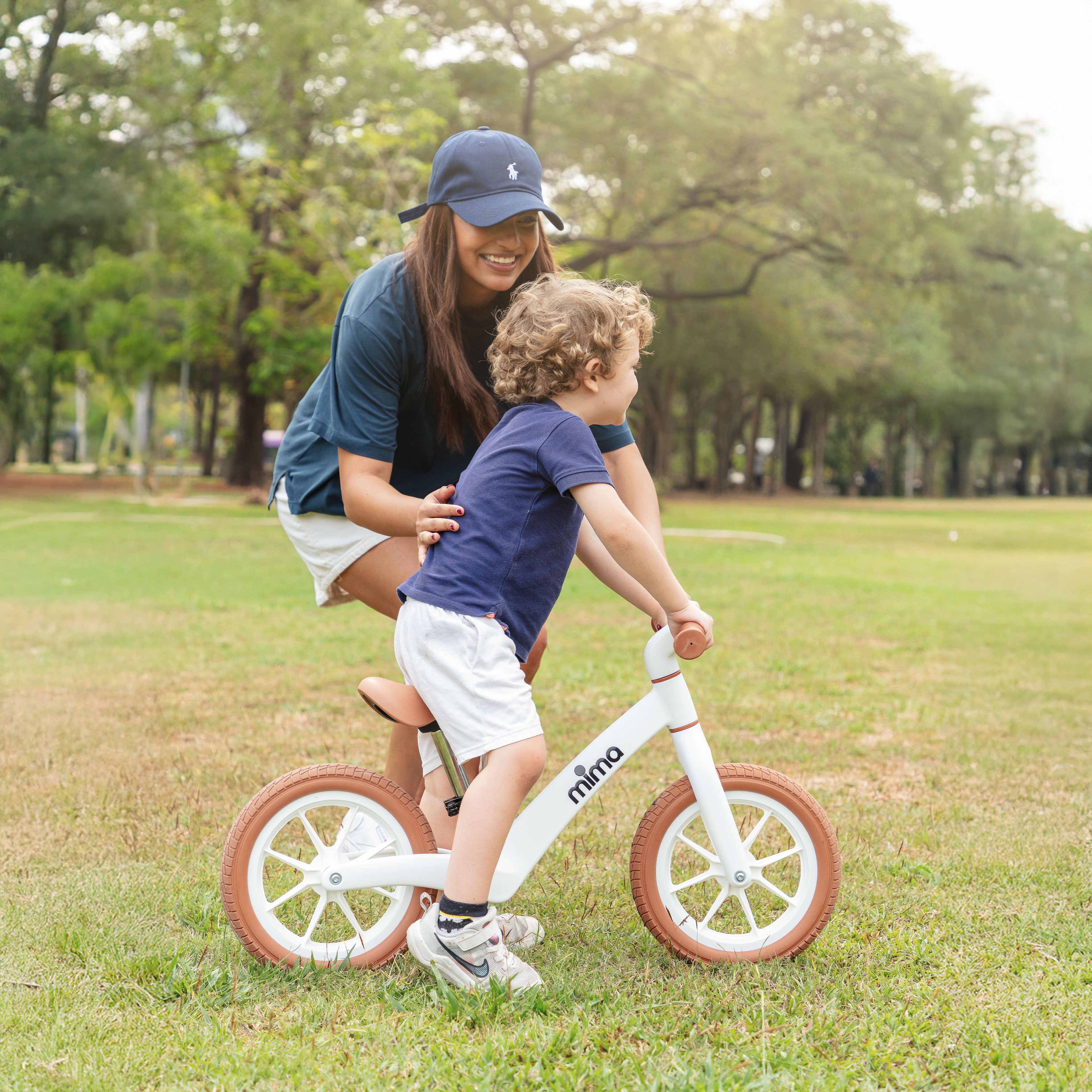 A smiling mother in a cap guiding her toddler riding a white mima zoom lite balance bike on a green grass field