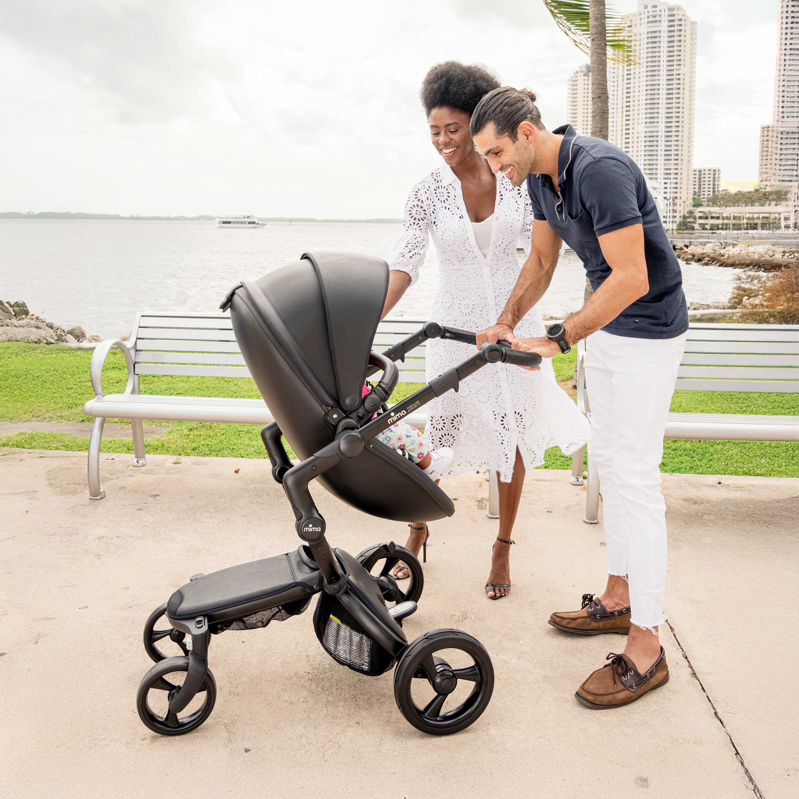 Mima Xari Black stroller on a dock with a waterfront view, featuring a couple with a city skyline and palm trees in the background, perfect for stylish family outings