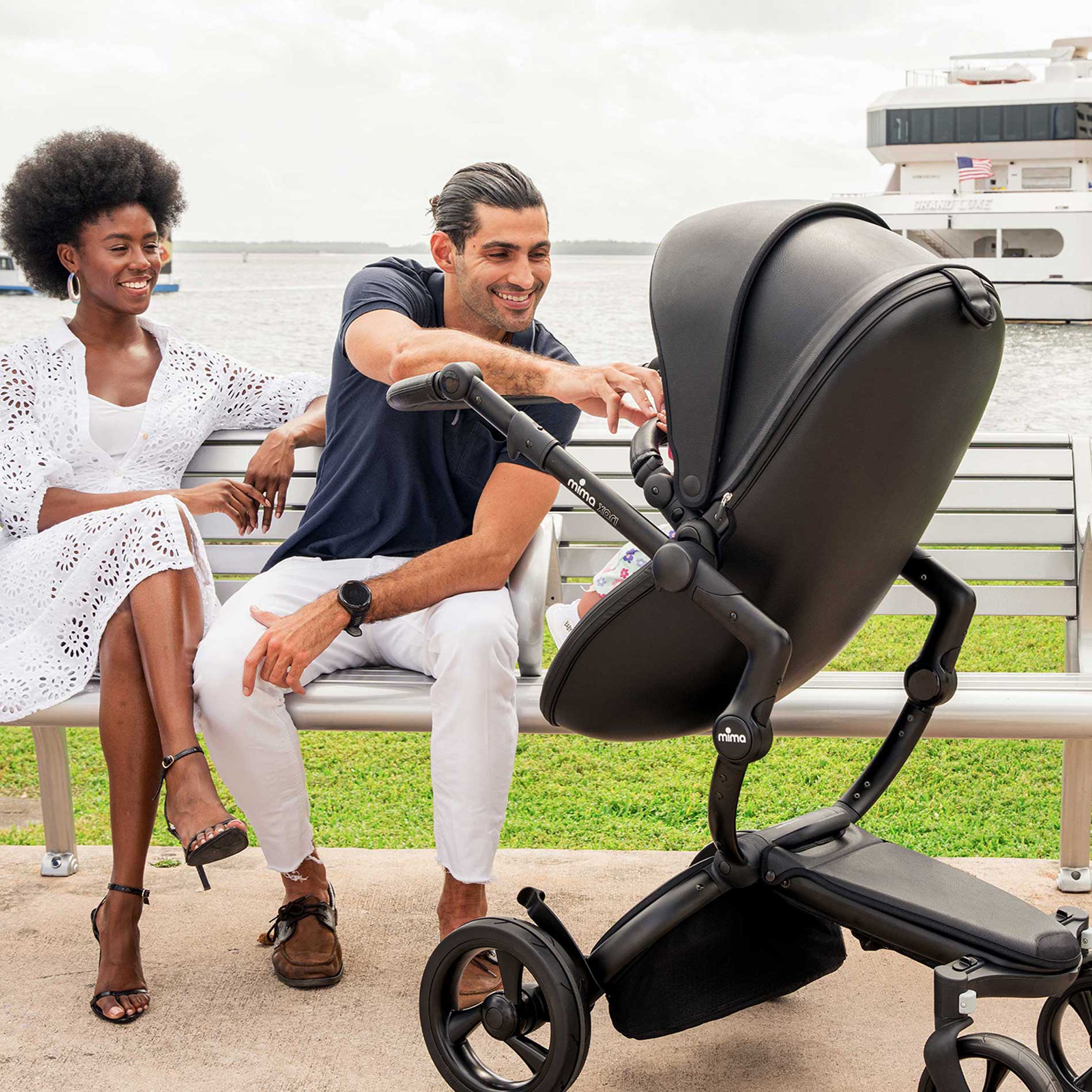 Mima Xari Black stroller near a dock with a couple sitting on a bench, ocean and a cruise ship in the background, surrounded by grassy area.