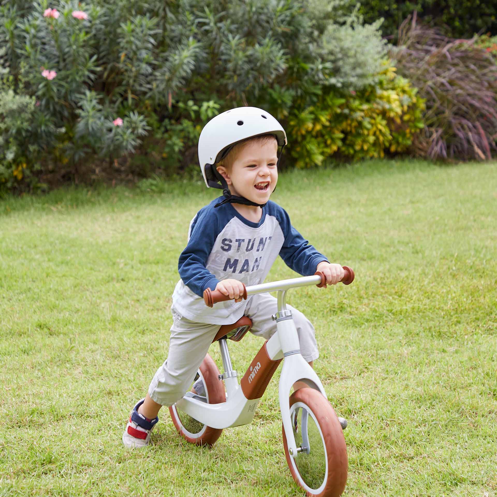 Boy wearing helmet, happily riding Mima® Zoom White balance bike on grassy field