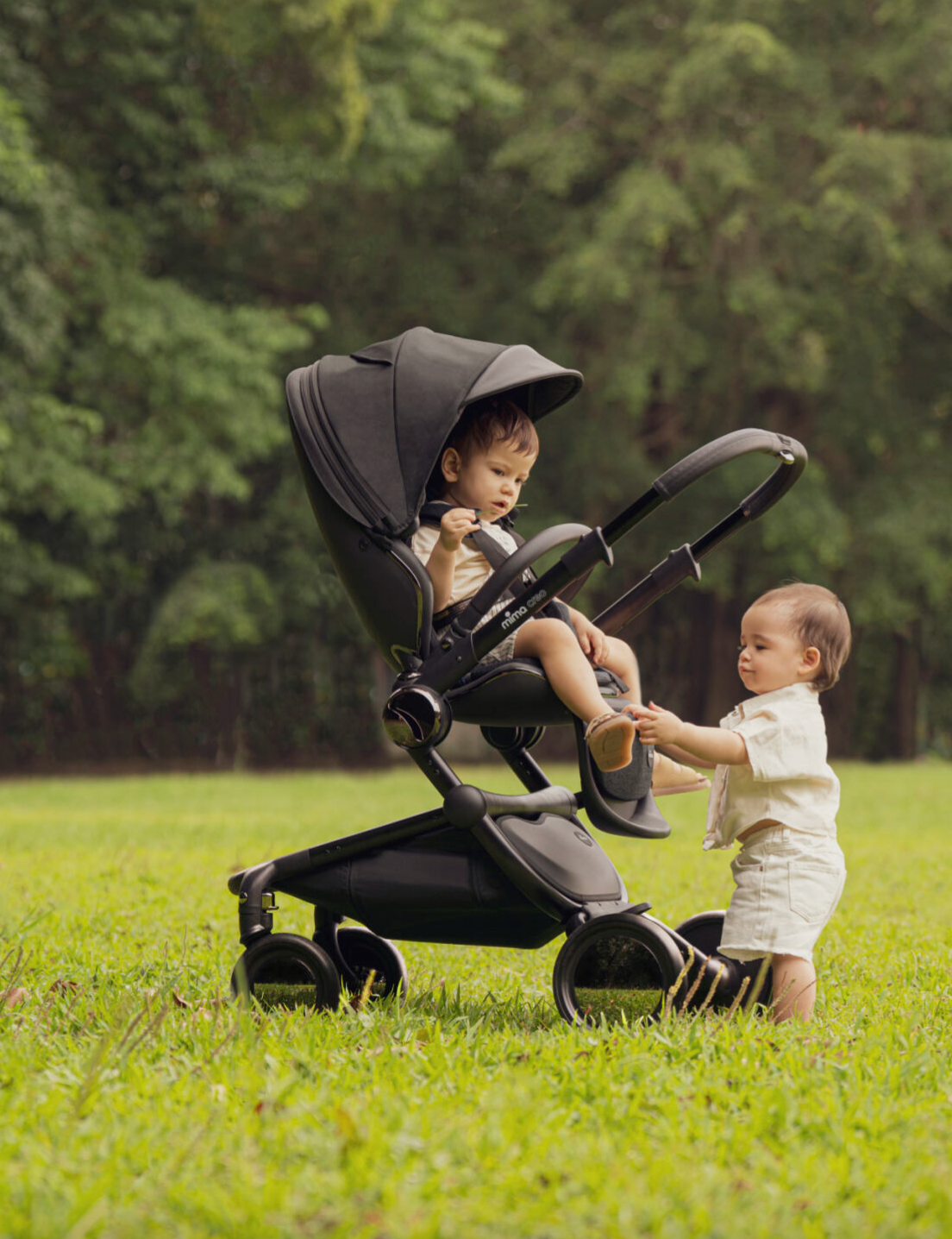 Mima Creo Black stroller in a park, featuring a child in the stroller and another toddler walking beside it, surrounded by lush green grass, ideal for family outings.