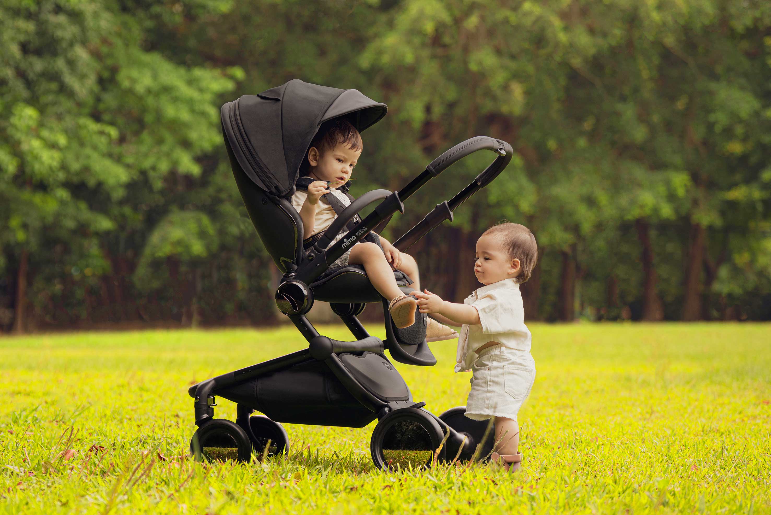 Mima Creo Black stroller in a park, featuring a child in the stroller and another toddler walking beside it, surrounded by lush green grass, ideal for family outings.