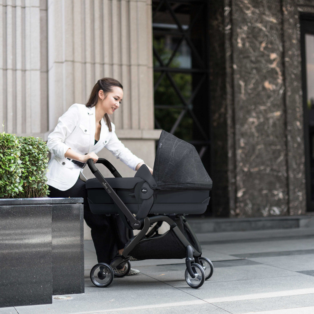 Mima® Zigi Bassinet in Ebony on a city street with a parent pushing, featuring a sleek black design and modern frame, ideal for urban baby transport.