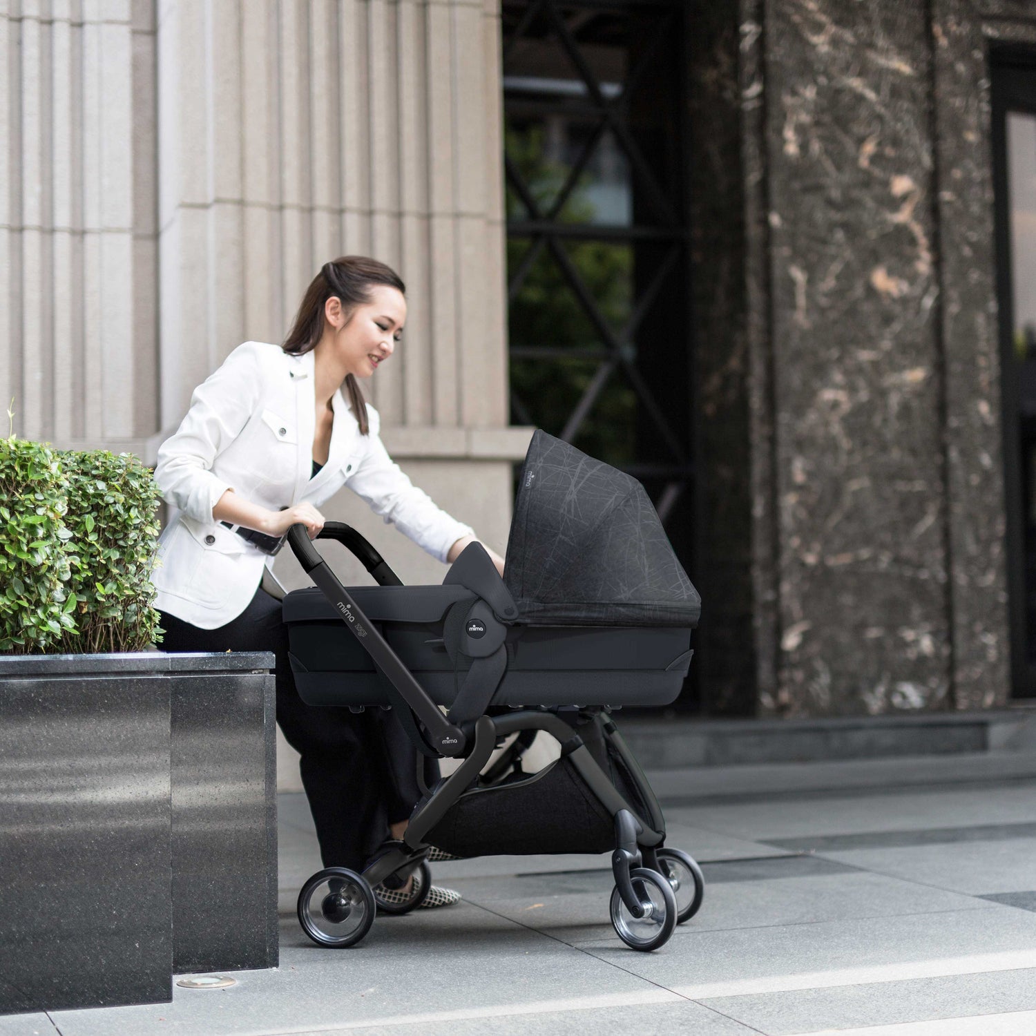 Mima® Zigi Bassinet in Ebony on a city street with a parent pushing, featuring a sleek black design and modern frame, ideal for urban baby transport.