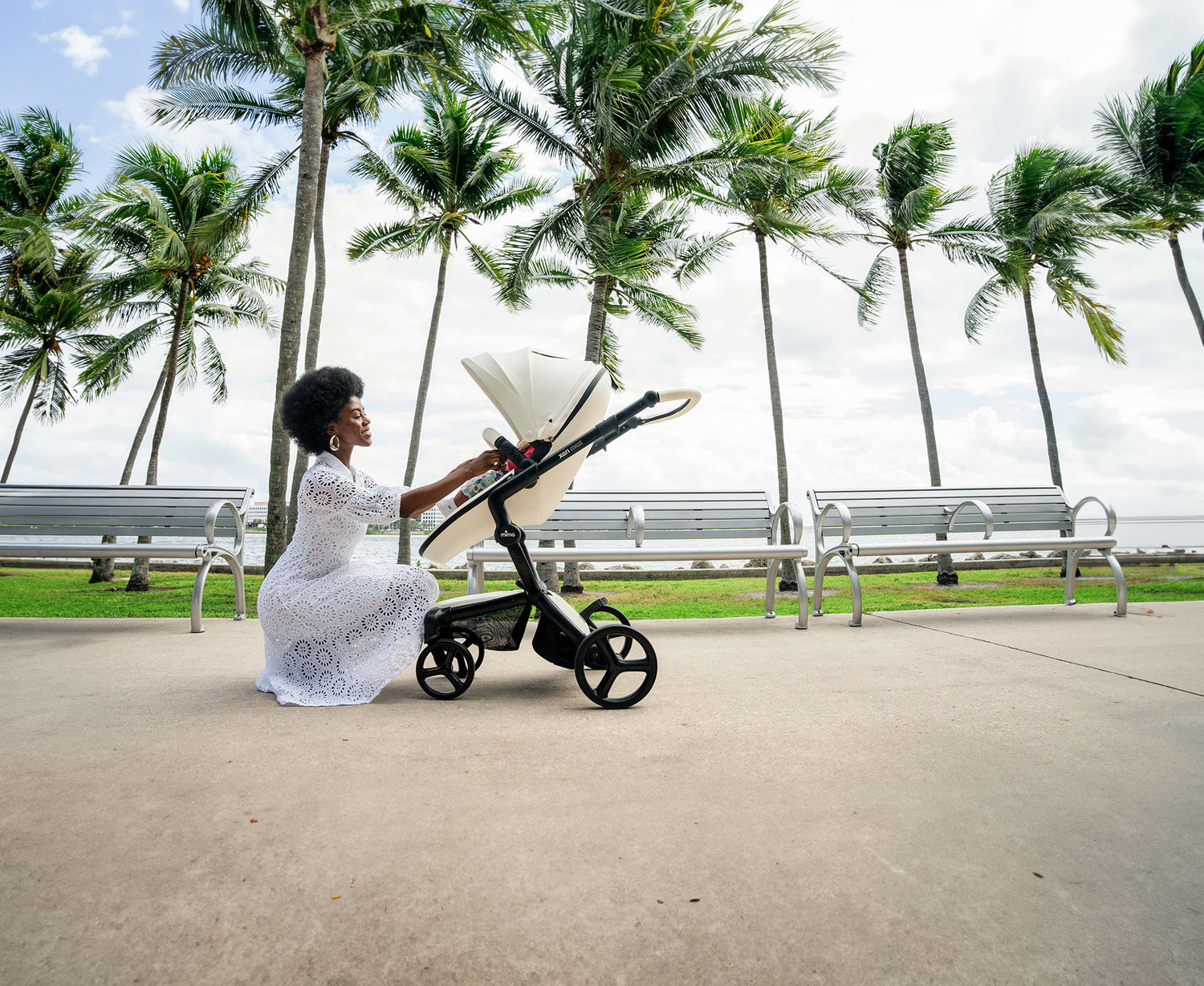 Xari Max Snow White stroller in a tropical setting with palm trees and ocean in the background, featuring a woman adjusting the stroller on a paved walkway near benches.
