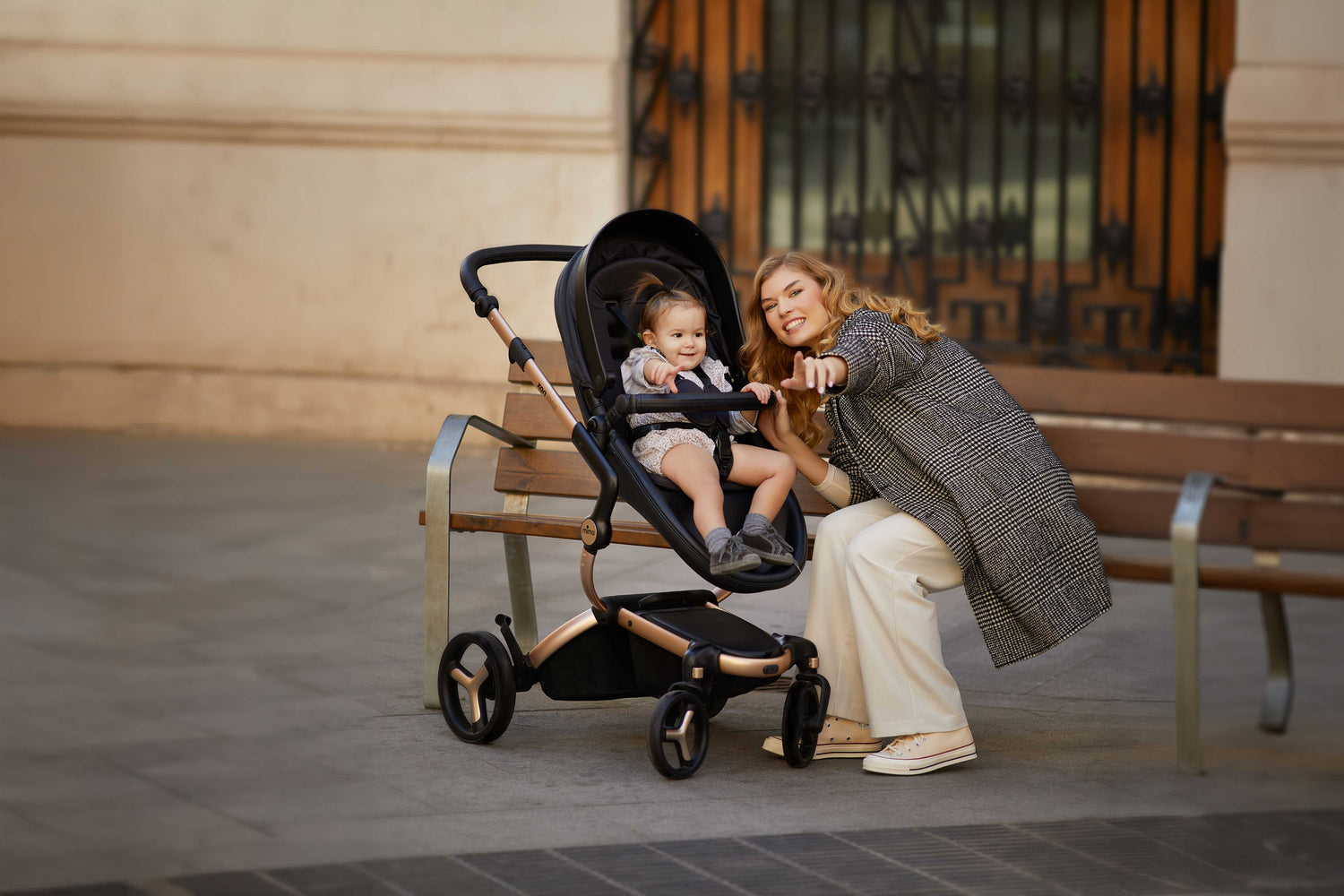 Mima® Xari Max Black Stroller on a city street, featuring a parent interacting with a child near a bench, showcasing its sleek design, ideal for stylish urban parenting.