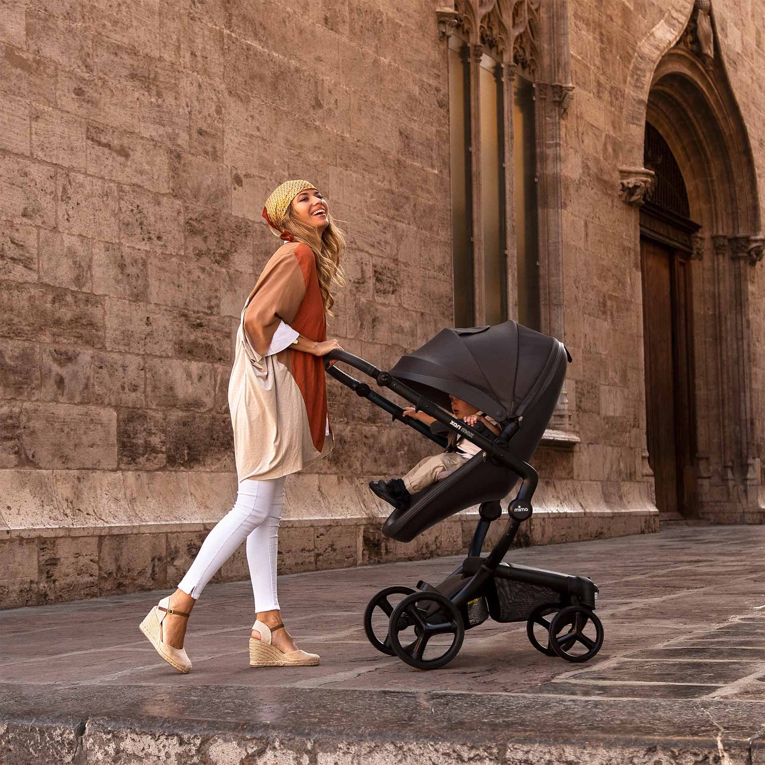 Black mima Xari Max stroller with baby, mother in beige hat pushing, side view on street