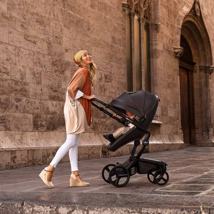 Black mima Xari Max stroller with baby, mother in beige hat pushing, side view on street