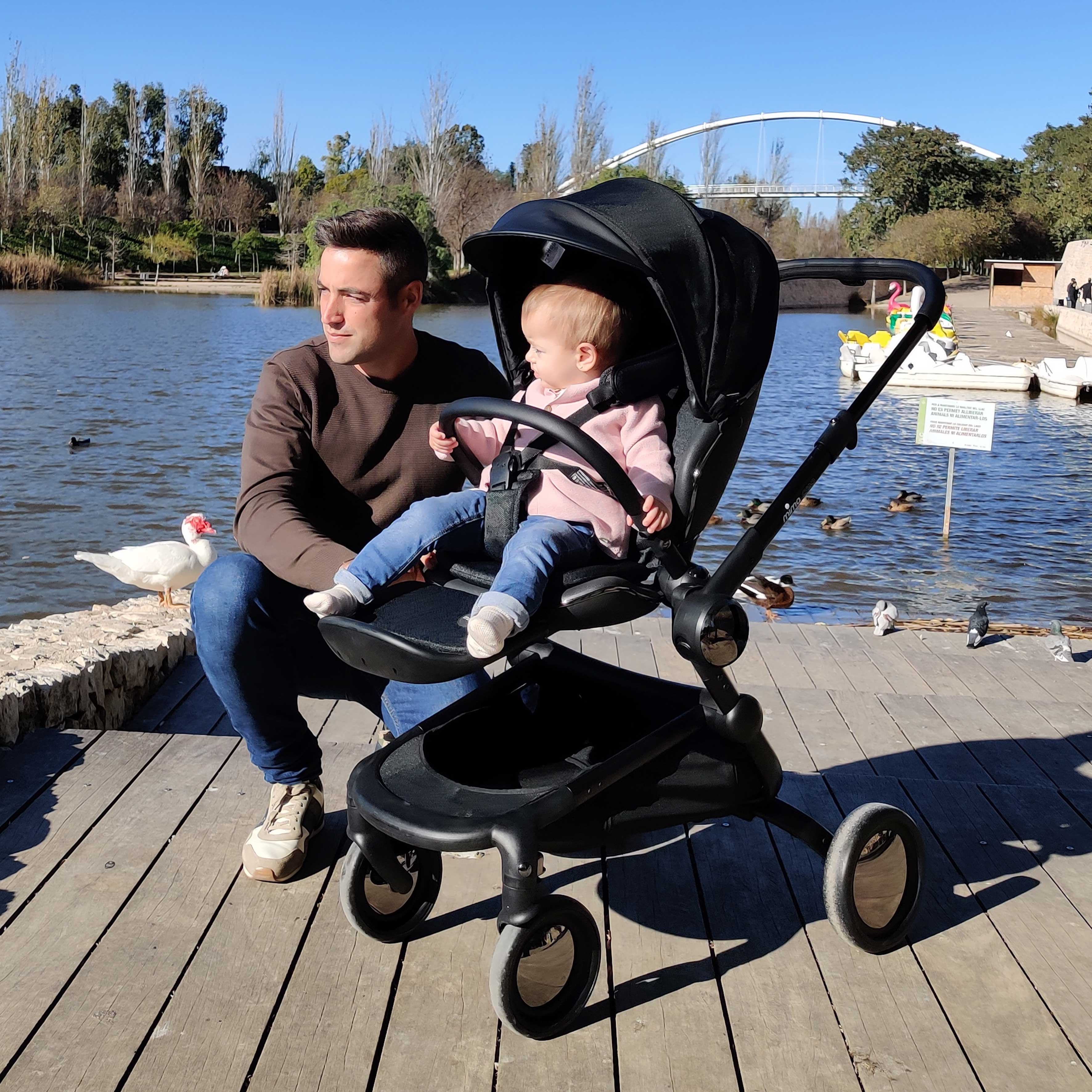 man kneeling next to toddler in mima creo compact stroller in black on a wooden deck near a lake 