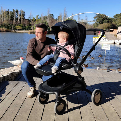 man kneeling next to toddler in mima creo compact stroller in black on a wooden deck near a lake 