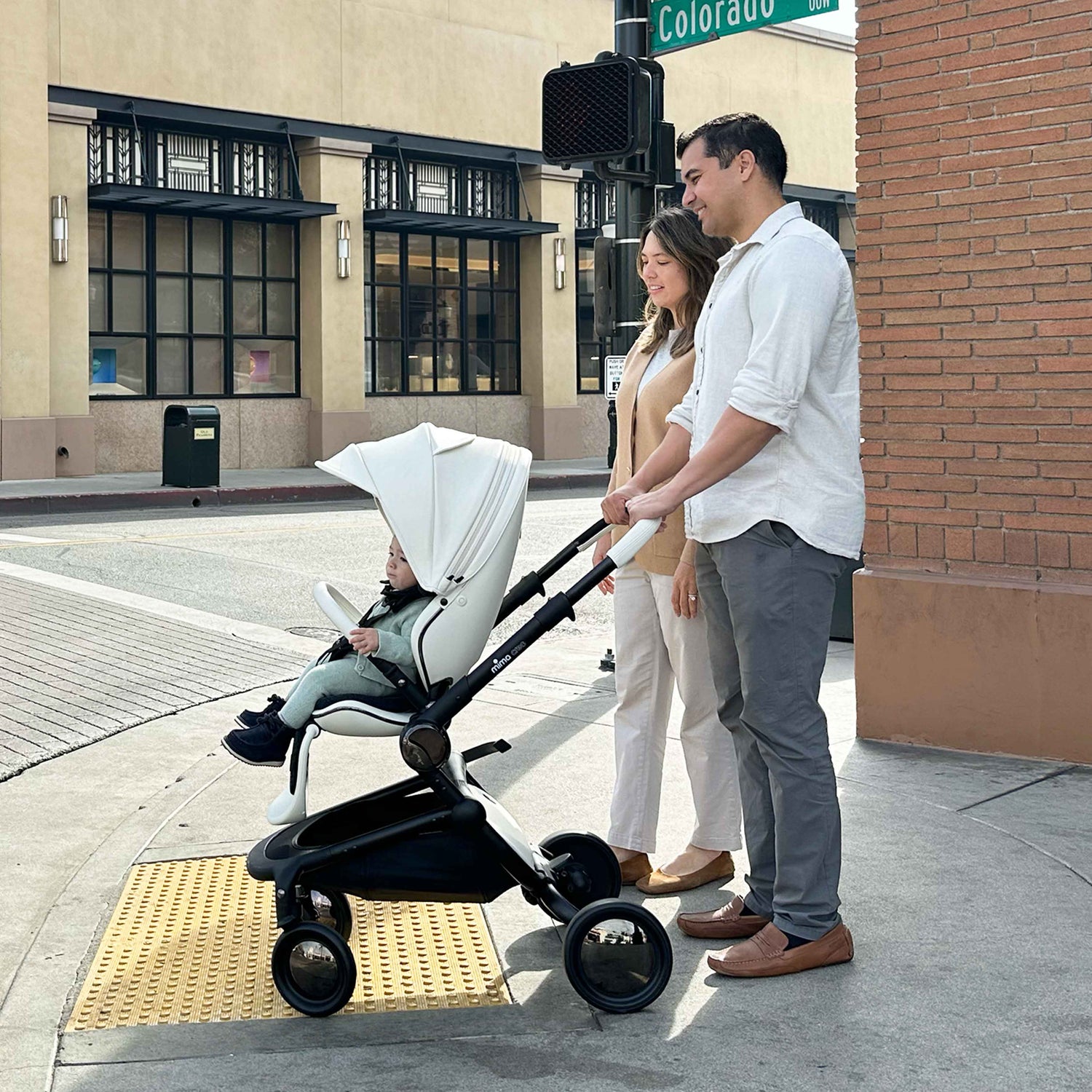 parents pushing child in mima creo stroller in white across a sunny city street intersection 