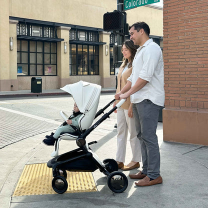 parents pushing child in mima creo stroller in white across a sunny city street intersection 