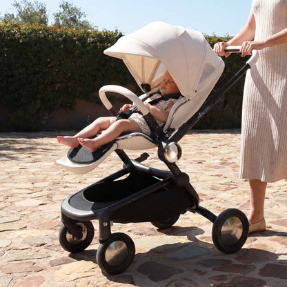 mima creo stroller in white, luxury baby stroller with white canopy, shown in an outdoor lifestyle setting with a woman walking on a stone walkway 