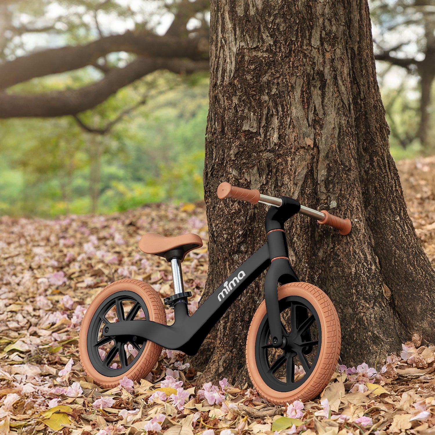 A sleek black mima zoom lite balance bike leaning against a rustic tree trunk in a park with fallen pink blossoms 