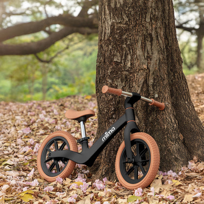 A sleek black mima zoom lite balance bike leaning against a rustic tree trunk in a park with fallen pink blossoms 