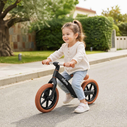 A toddler riding a black mima zoom lite balance bike on an outdoor paved surface 