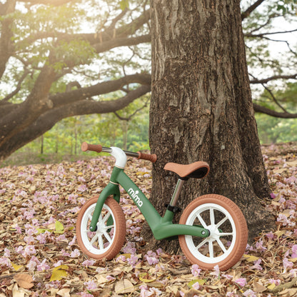 A green mima zoom lite balance bike leaning against a large tree trunk on a ground covered with fallen autumn leaves 