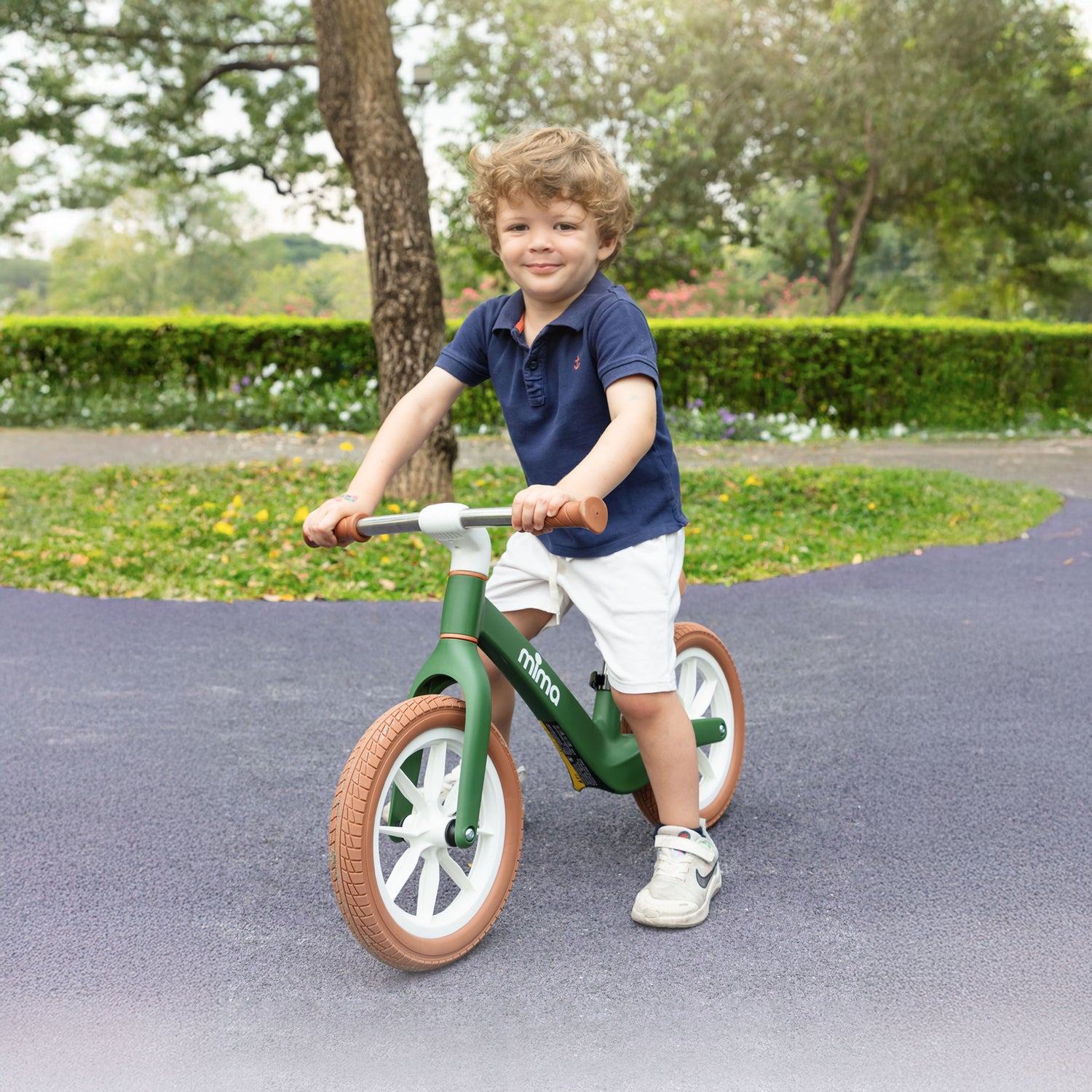 A toddler in a navy polo shirt riding a green mima zoom lite balance bike on a paved park pathway 