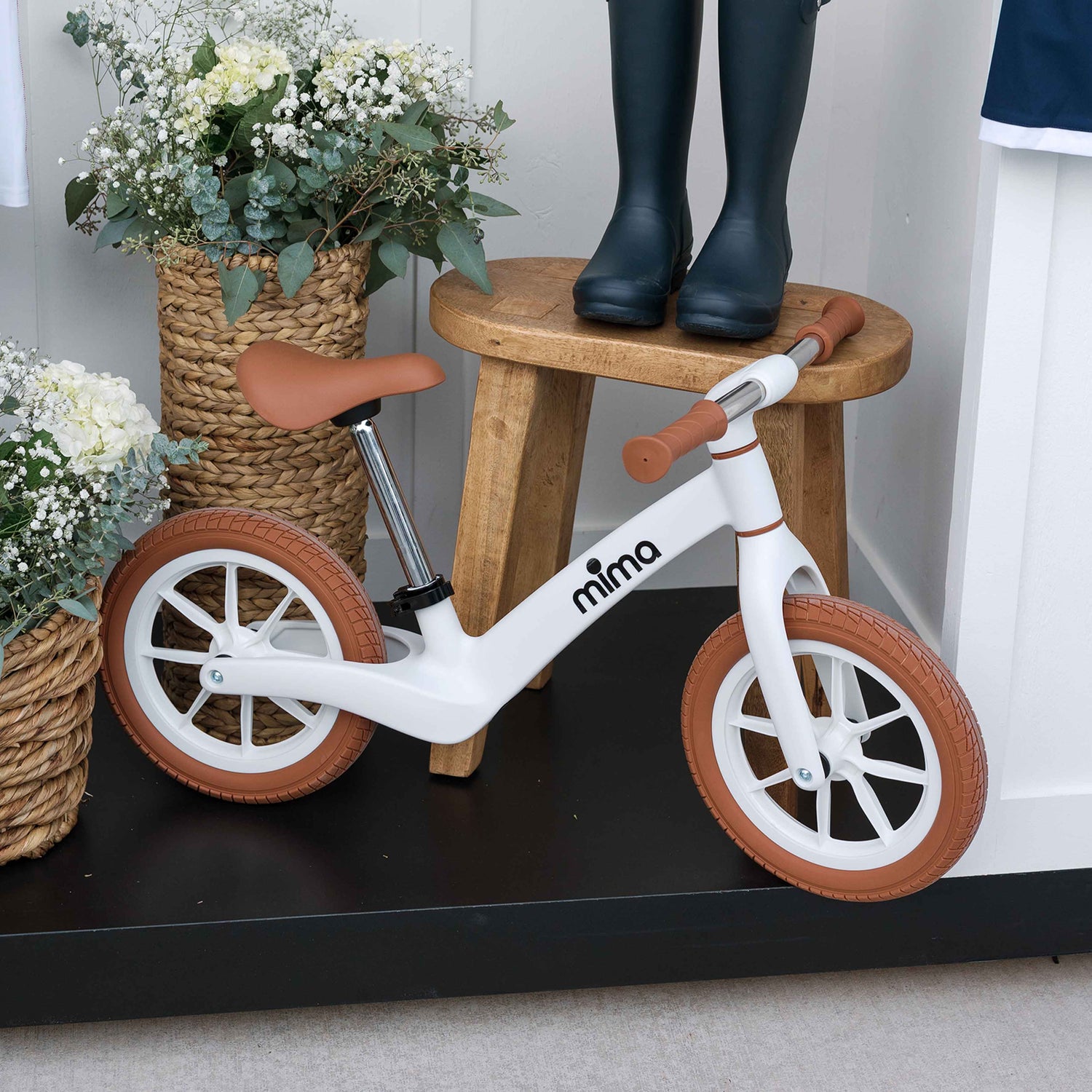 A white mima zoom lite balance bike displayed indoors next to a wooden stool, rain boots, and a basket of flowers 