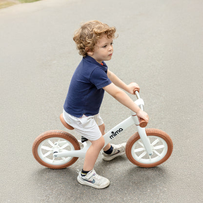 A toddler with curly hair wearing a navy polo shirt riding a white mima zoom lite balance bike on an asphalt road 